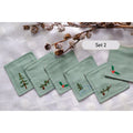 Set of six green linen coasters with embroidered winter designs, including a pine tree and a holly sprig — hand-embroidered by The Thread Nest