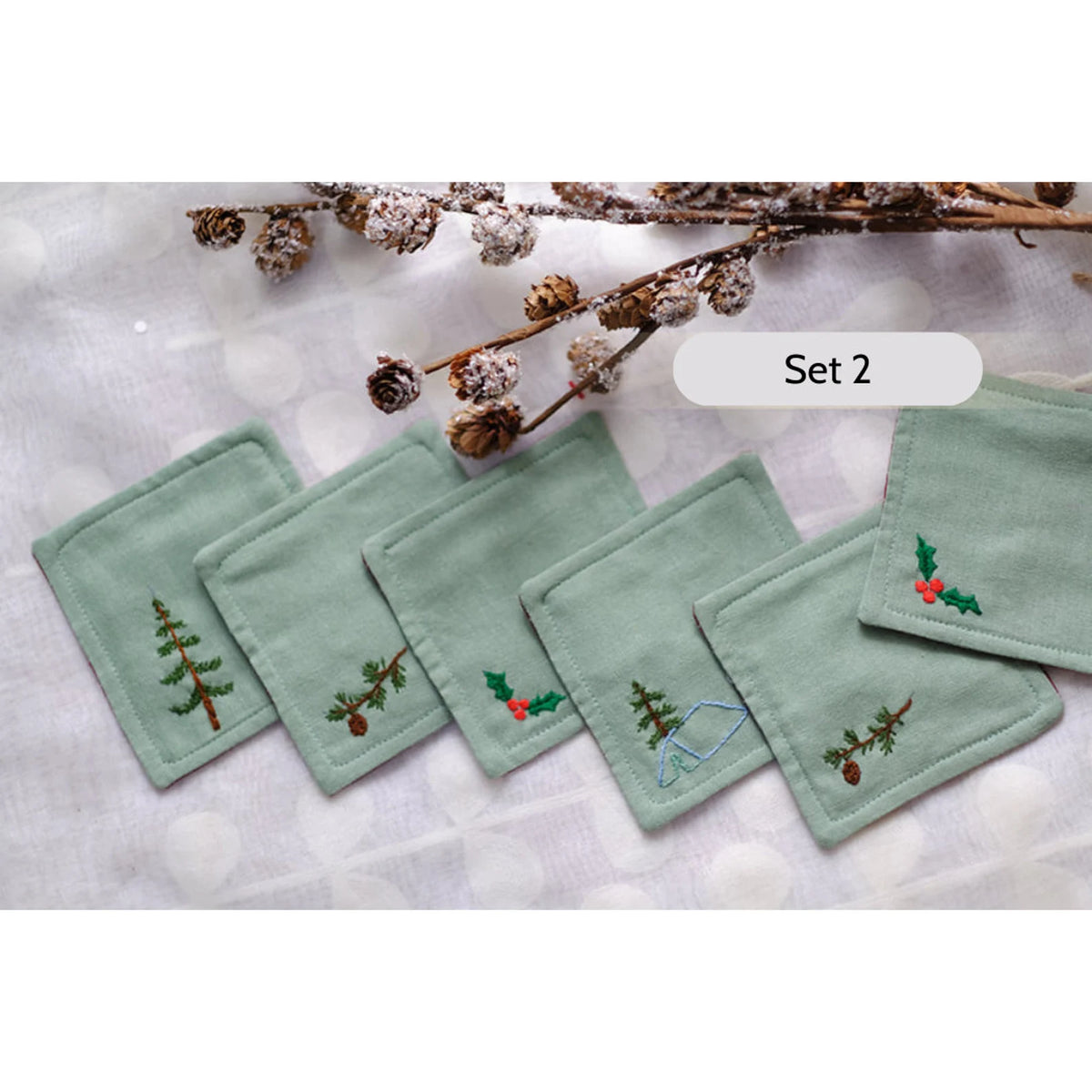 Set of six green linen coasters with embroidered winter designs, including a pine tree and a holly sprig — hand-embroidered by The Thread Nest