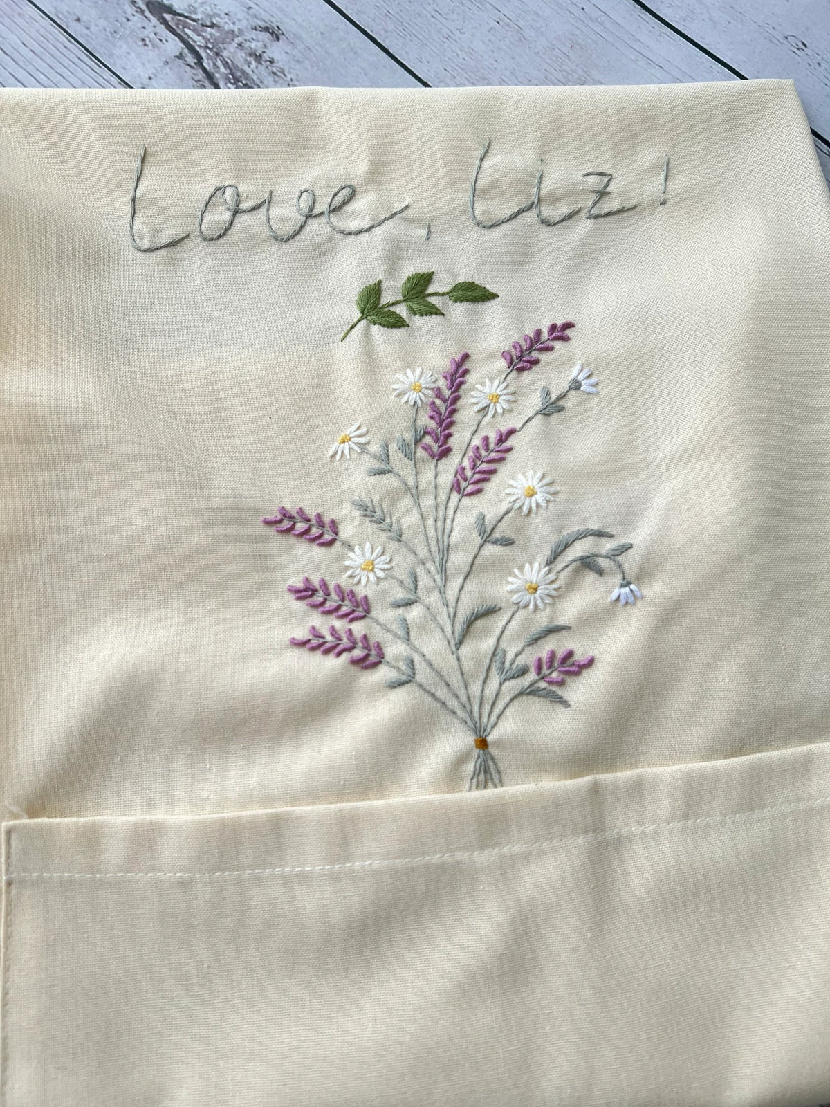Close-up of a cream linen apron embroidered with lavender and the words Love, Liz — hand-embroidered by The Thread Nest