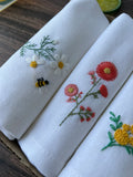 Three white linen napkins with embroidered floral designs — hand-embroidered by The Thread Nest