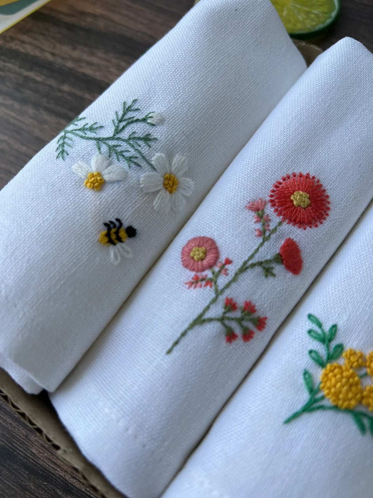 Three white linen napkins with embroidered floral designs — hand-embroidered by The Thread Nest