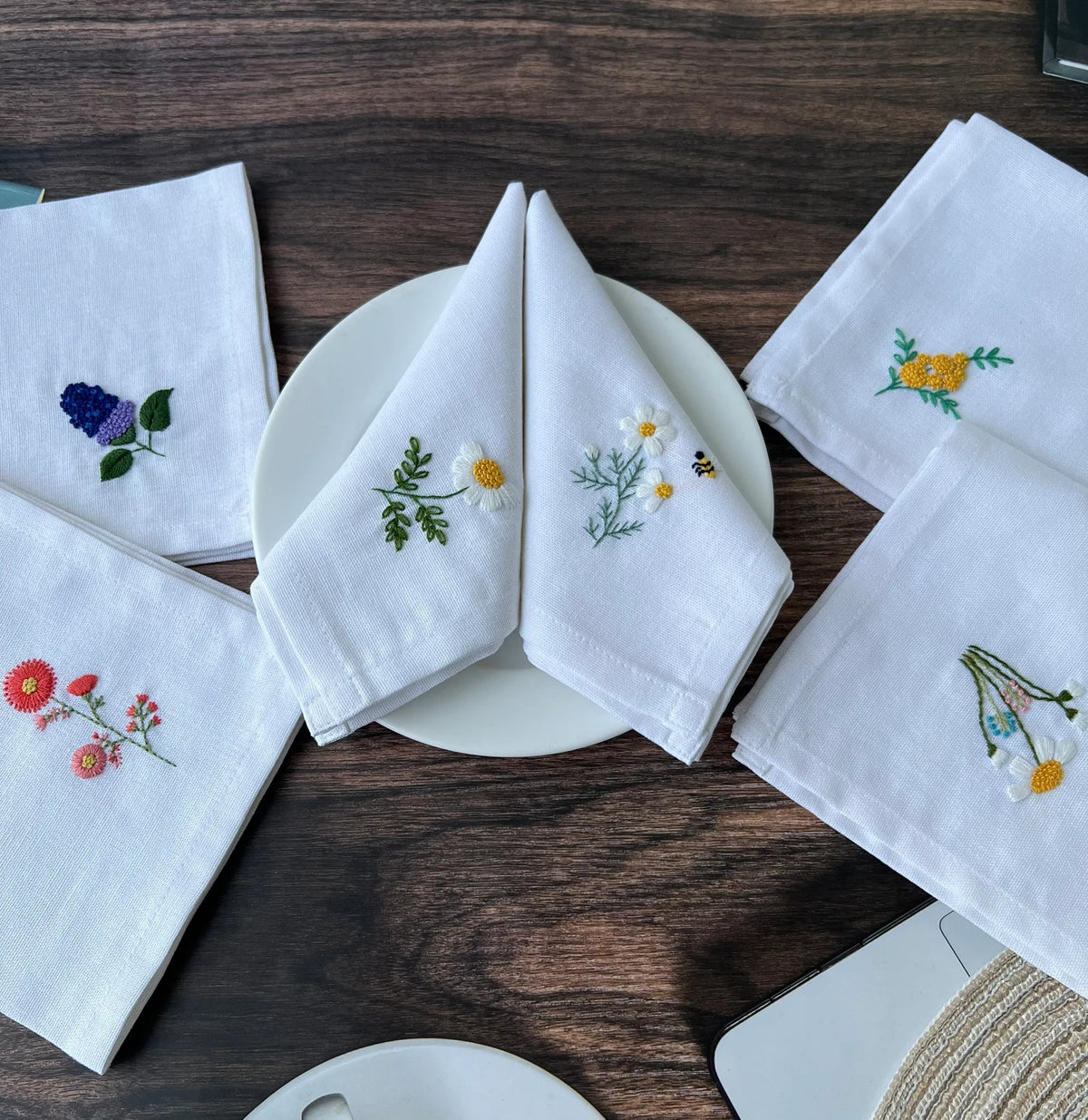 Five white linen napkins with embroidered floral designs — hand-embroidered by The Thread Nest
