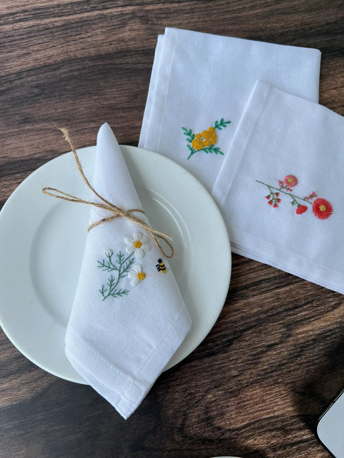 Three white linen napkins with embroidered floral designs — hand-embroidered by The Thread Nest