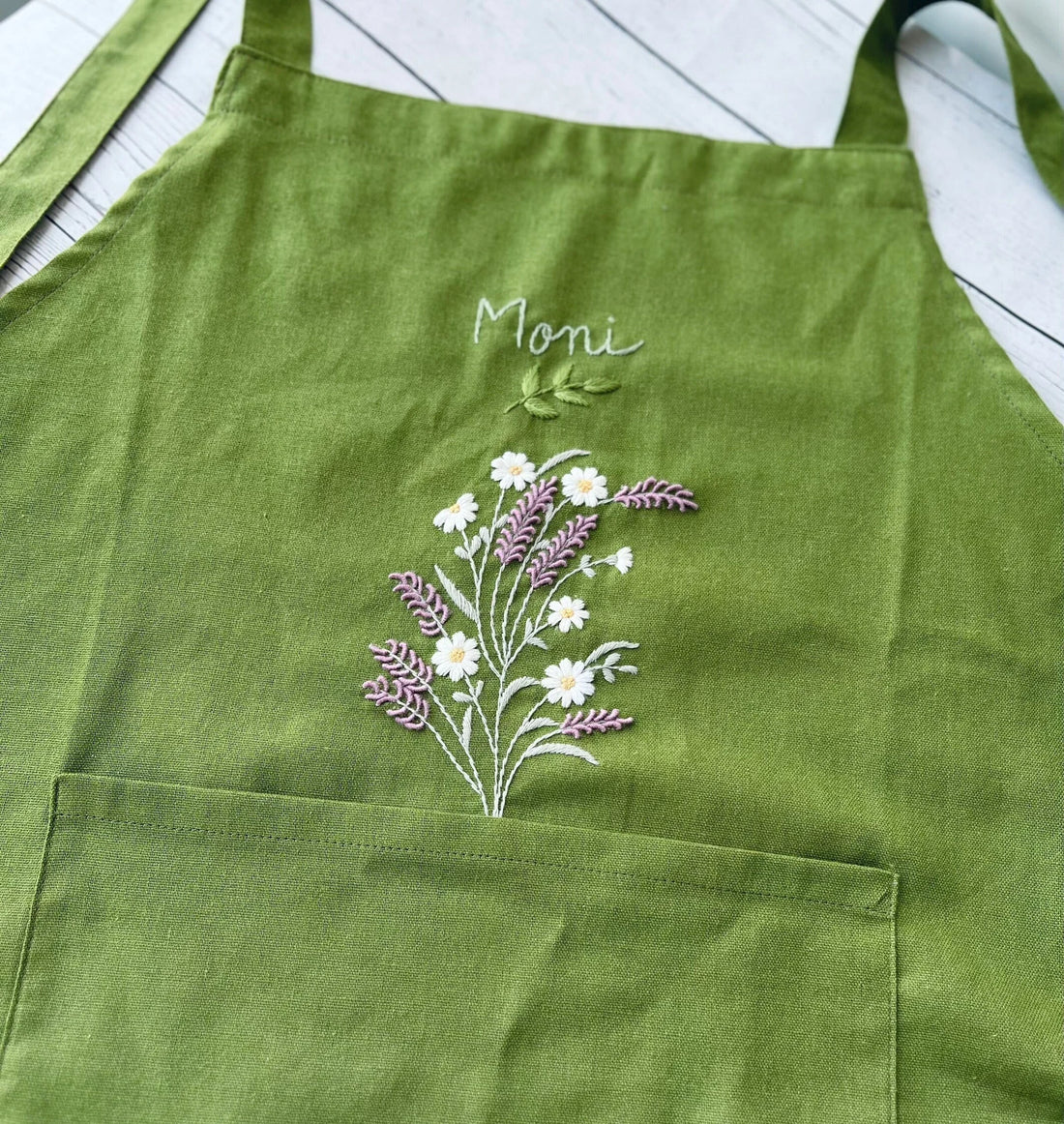 A woman wearing a green embroidered linen apron, whisking a bowl in a kitchen — hand-embroidered by The Thread Nest