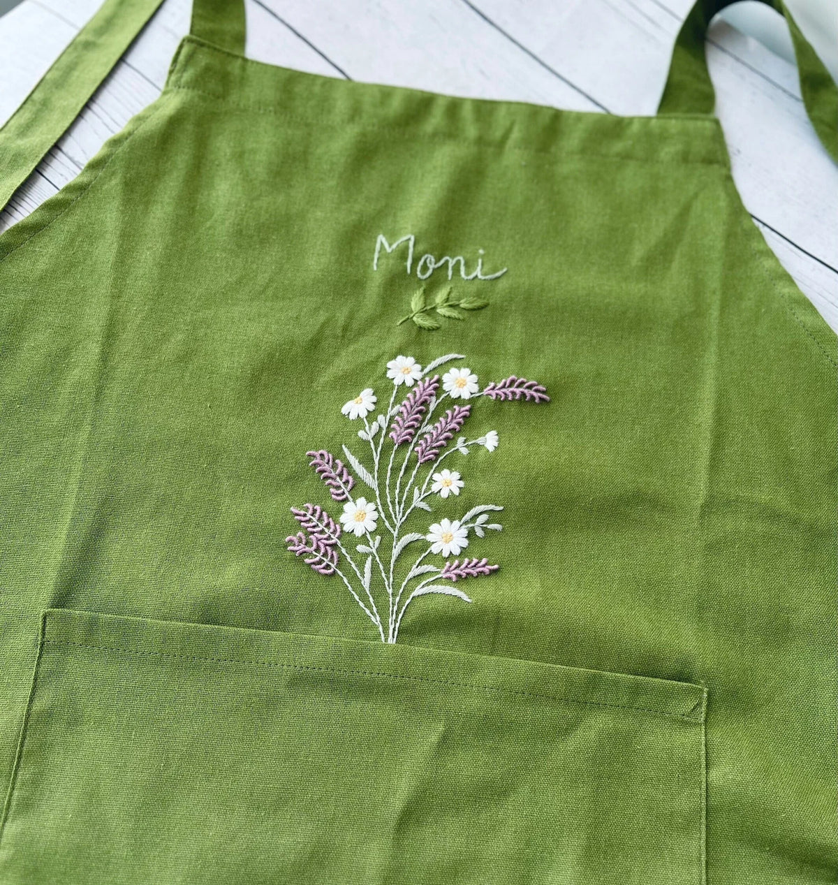 Close-up of a green linen apron bib embroidered with a lavender and daisy bouquet and the name Moni — hand-embroidered by The Thread Nest