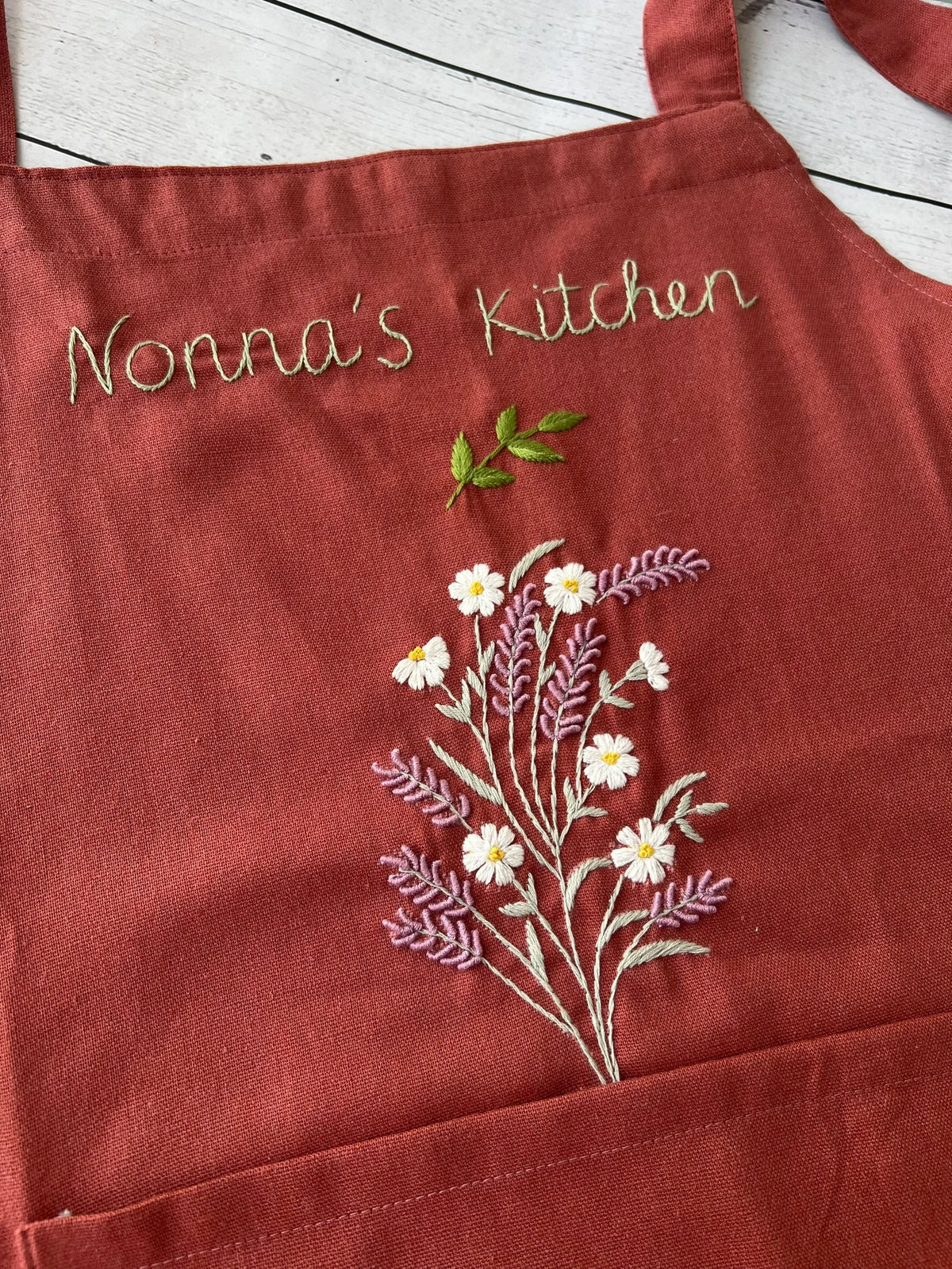 A rust linen apron embroidered with a flower bouquet and the words Nonna's Kitchen — hand-embroidered by The Thread Nest