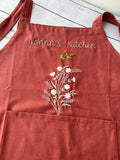 A rust linen apron with a front pocket, embroidered with flowers and the words Nonna's Kitchen — hand-embroidered by The Thread Nest