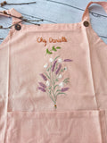 A pink linen apron with wooden buttons on the straps, embroidered with lavender and a name — hand-embroidered by The Thread Nest