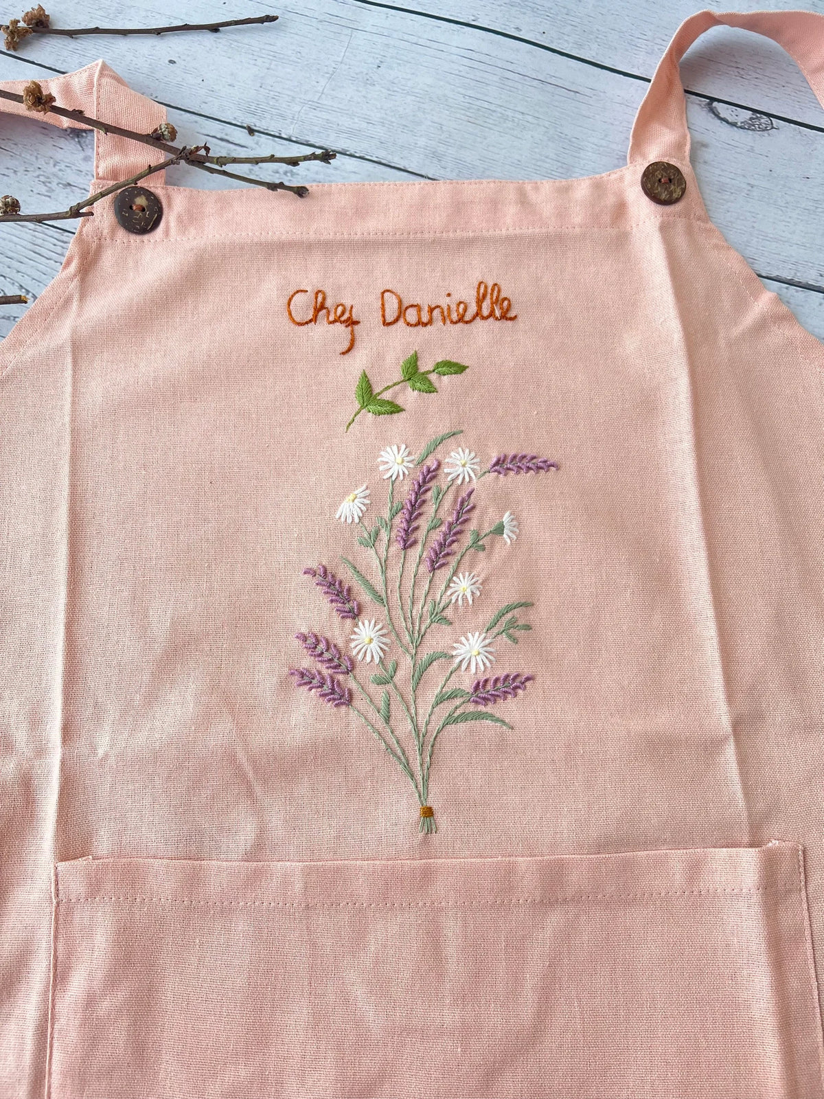 A pink linen apron with wooden buttons on the straps, embroidered with lavender and a name — hand-embroidered by The Thread Nest