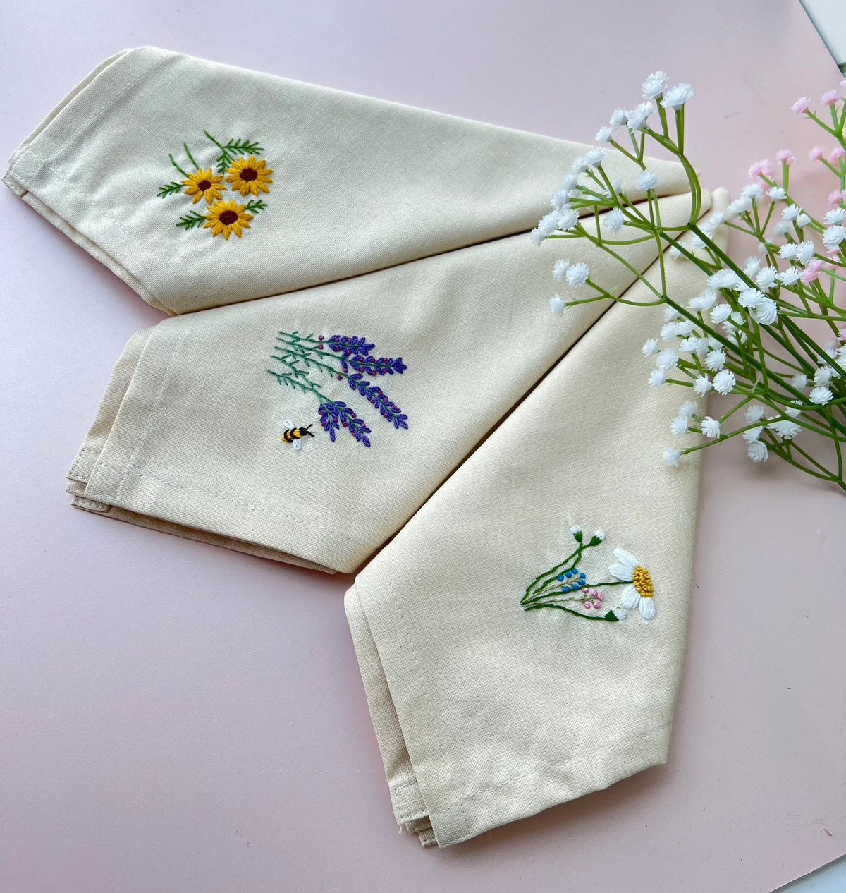 Three white linen tea towels with embroidered floral designs — hand-embroidered by The Thread Nest