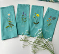 Four teal blue linen napkins with embroidered floral designs and a stitched name — hand-embroidered by The Thread Nest