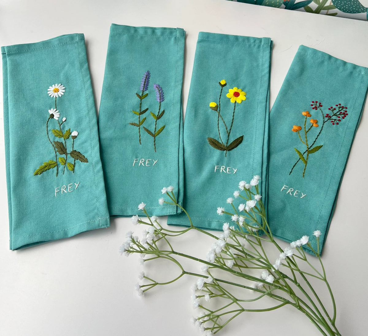 Four teal blue linen napkins with embroidered floral designs and a stitched name — hand-embroidered by The Thread Nest