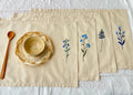A set of beige linen placemats with embroidered floral designs — hand-embroidered by The Thread Nest