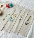 Four beige linen placemats with embroidered floral designs — hand-embroidered by The Thread Nest