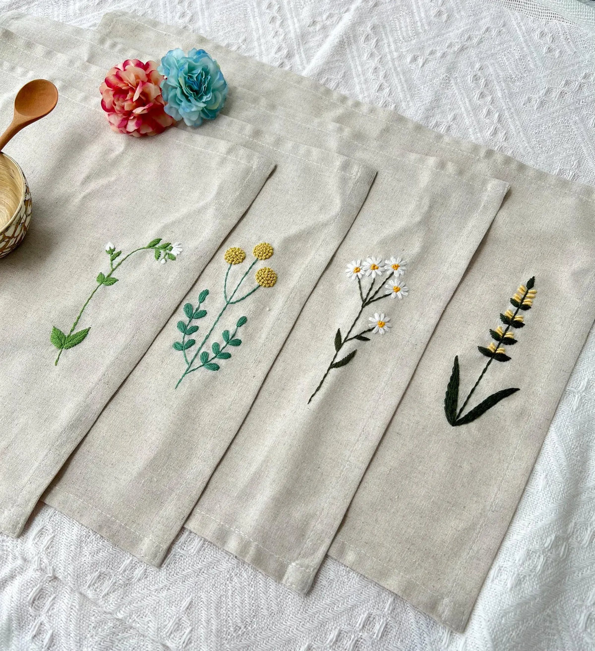Four beige linen placemats with embroidered floral designs — hand-embroidered by The Thread Nest
