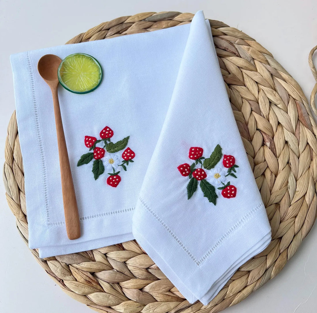 A white linen napkin with embroidered strawberries and a white flower — hand-embroidered by The Thread Nest