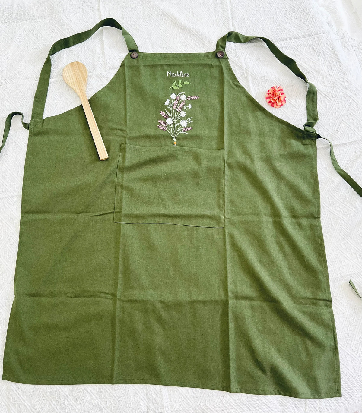 An olive green linen apron laid flat with a front pocket and an embroidered name — hand-embroidered by The Thread Nest