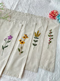 Five beige linen napkins with embroidered floral designs — hand-embroidered by The Thread Nest