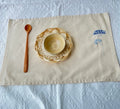 A beige linen placemat set with a woven wicker plate and a small wooden bowl — hand-embroidered by The Thread Nest