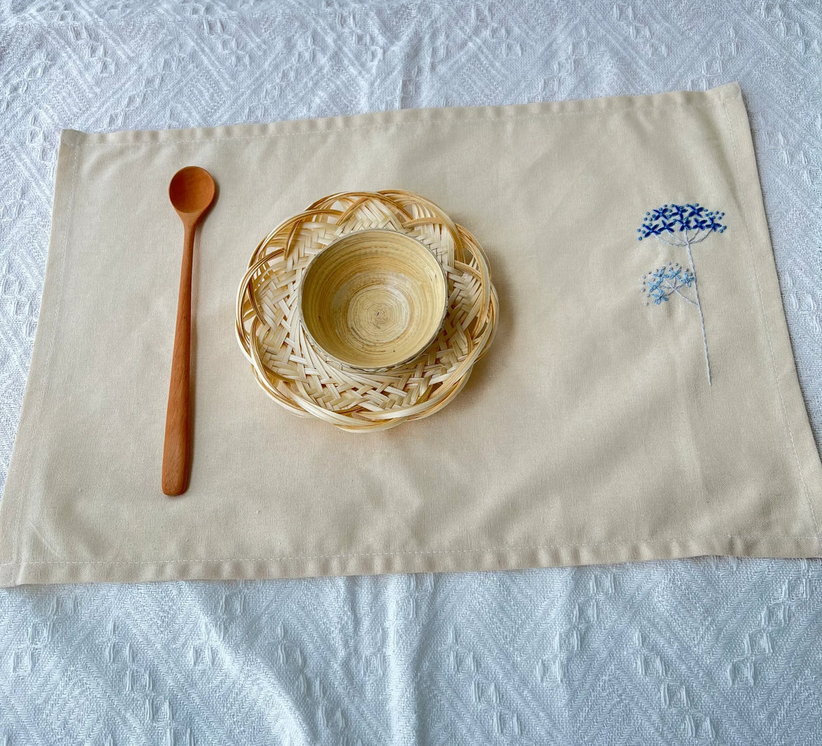A beige linen placemat set with a woven wicker plate and a small wooden bowl — hand-embroidered by The Thread Nest