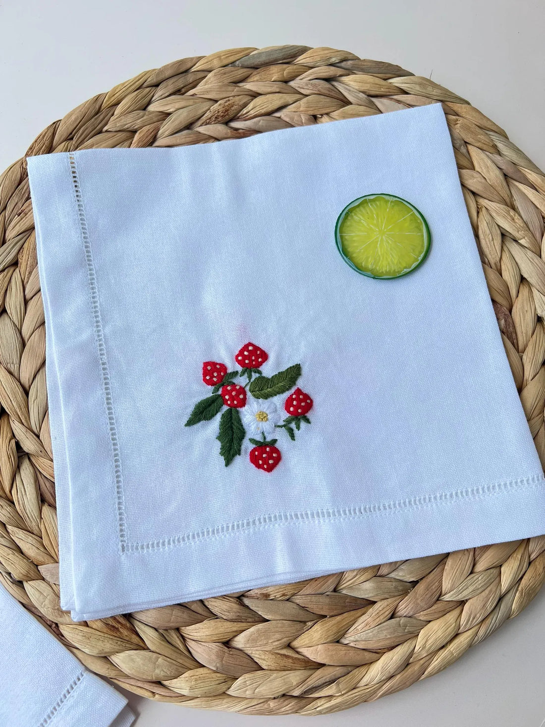 A white linen napkin with embroidered strawberries and a white flower — hand-embroidered by The Thread Nest