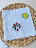 A white linen napkin with embroidered strawberries and a white flower — hand-embroidered by The Thread Nest