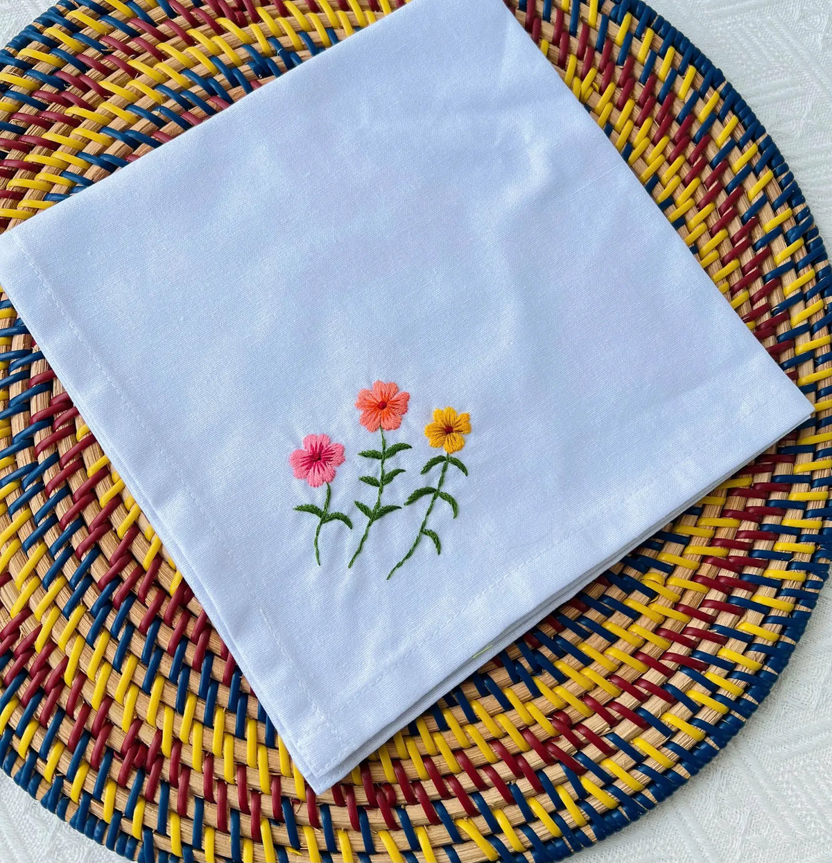 A white linen napkin with embroidered pink, orange and yellow flowers — hand-embroidered by The Thread Nest
