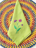 A green linen napkin with embroidered purple flowers — hand-embroidered by The Thread Nest