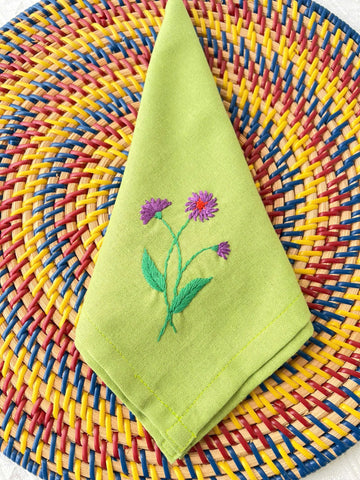 A green linen napkin with embroidered purple flowers — hand-embroidered by The Thread Nest