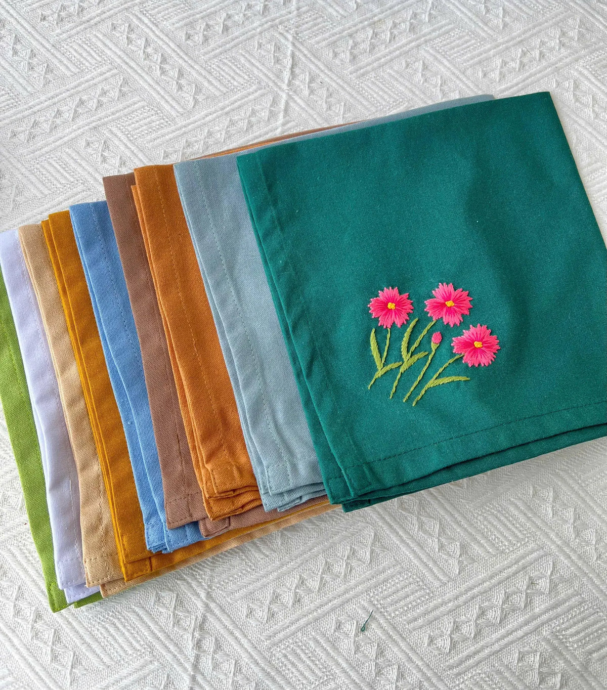 A stack of ten embroidered napkins in green, blue, brown, yellow and beige — hand-embroidered by The Thread Nest