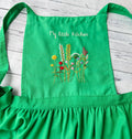 A green linen apron embroidered with wildflowers and the words My Little Kitchen — hand-embroidered by The Thread Nest