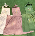 Three embroidered linen aprons in cream, pink and green laid out side by side — hand-embroidered by The Thread Nest