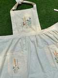 A cream linen apron with a front pocket, embroidered with wildflowers, laid on grass — hand-embroidered by The Thread Nest