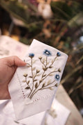 A white linen handkerchief with embroidered blue flowers and brown stems — hand-embroidered by The Thread Nest