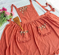 A rust ruffled linen apron laid flat with a wooden spoon and pink flowers — hand-embroidered by The Thread Nest