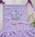 A lavender linen kids apron with a ruffled hem, embroidered with daisies, lavender and bees — hand-embroidered by The Thread Nest