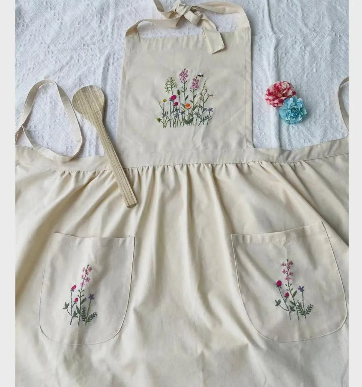 A cream linen kids apron laid flat with embroidered wildflowers on the bib and pockets — hand-embroidered by The Thread Nest