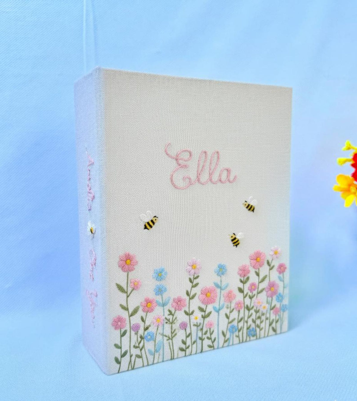A light beige photo album with a name in pink and embroidered flowers — hand-embroidered by The Thread Nest
