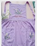 A lavender linen kids apron with two pockets, embroidered with wildflowers and a name — hand-embroidered by The Thread Nest