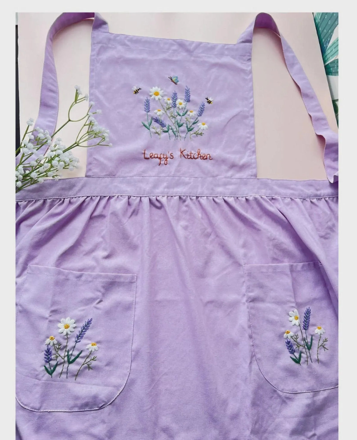 A lavender linen kids apron with two pockets, embroidered with wildflowers and a name — hand-embroidered by The Thread Nest