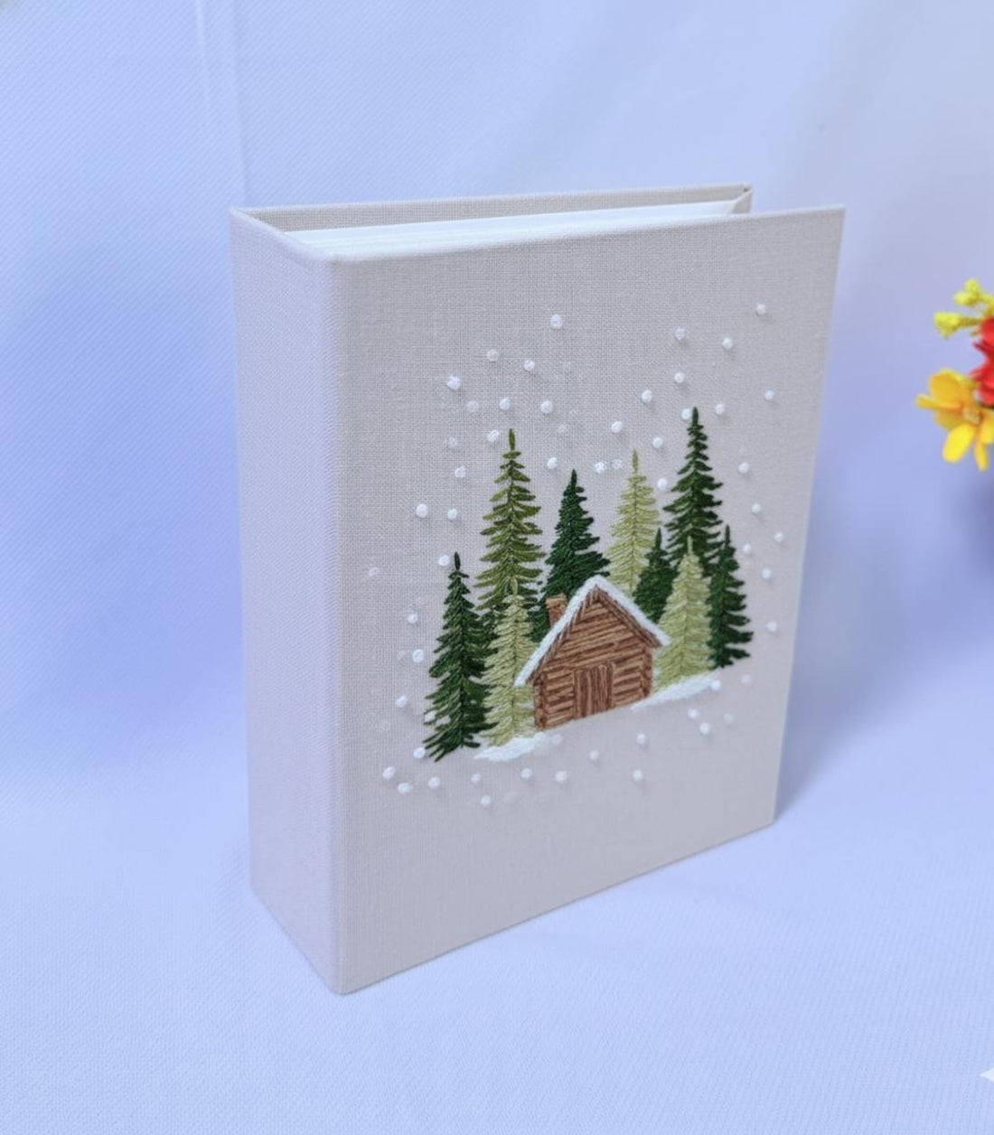 A beige book with an embroidered winter scene of a cabin and pine trees — hand-embroidered by The Thread Nest