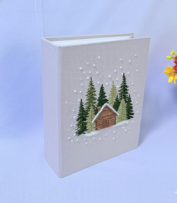 A beige book with an embroidered winter scene of a cabin and pine trees — hand-embroidered by The Thread Nest
