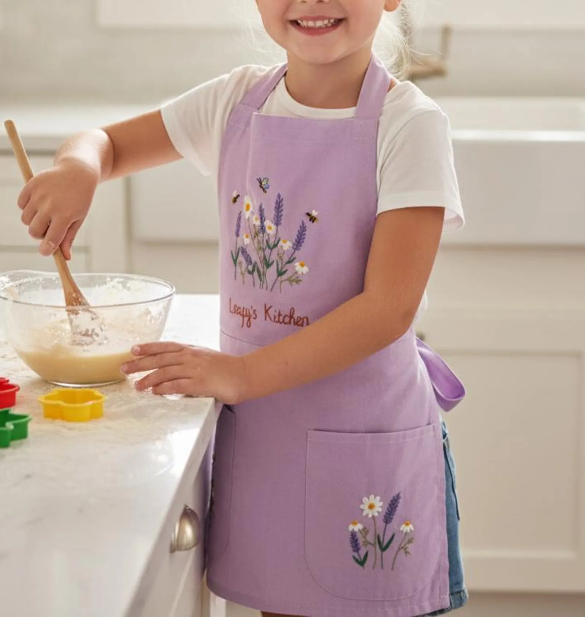 A child in a lavender embroidered linen apron stirring a bowl — hand-embroidered by The Thread Nest