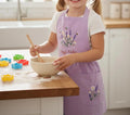 A child wearing a lavender embroidered linen apron, mixing a bowl at a kitchen counter — hand-embroidered by The Thread Nest