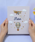 A light gray book with an embroidered yellow biplane and a hot air balloon — hand-embroidered by The Thread Nest