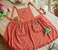 A rust linen ruffled apron laid flat, embroidered with white flowers on the bib and pockets — hand-embroidered by The Thread Nest