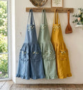 Three ruffled linen aprons in blue, green and mustard hanging on a wooden rack — hand-embroidered by The Thread Nest