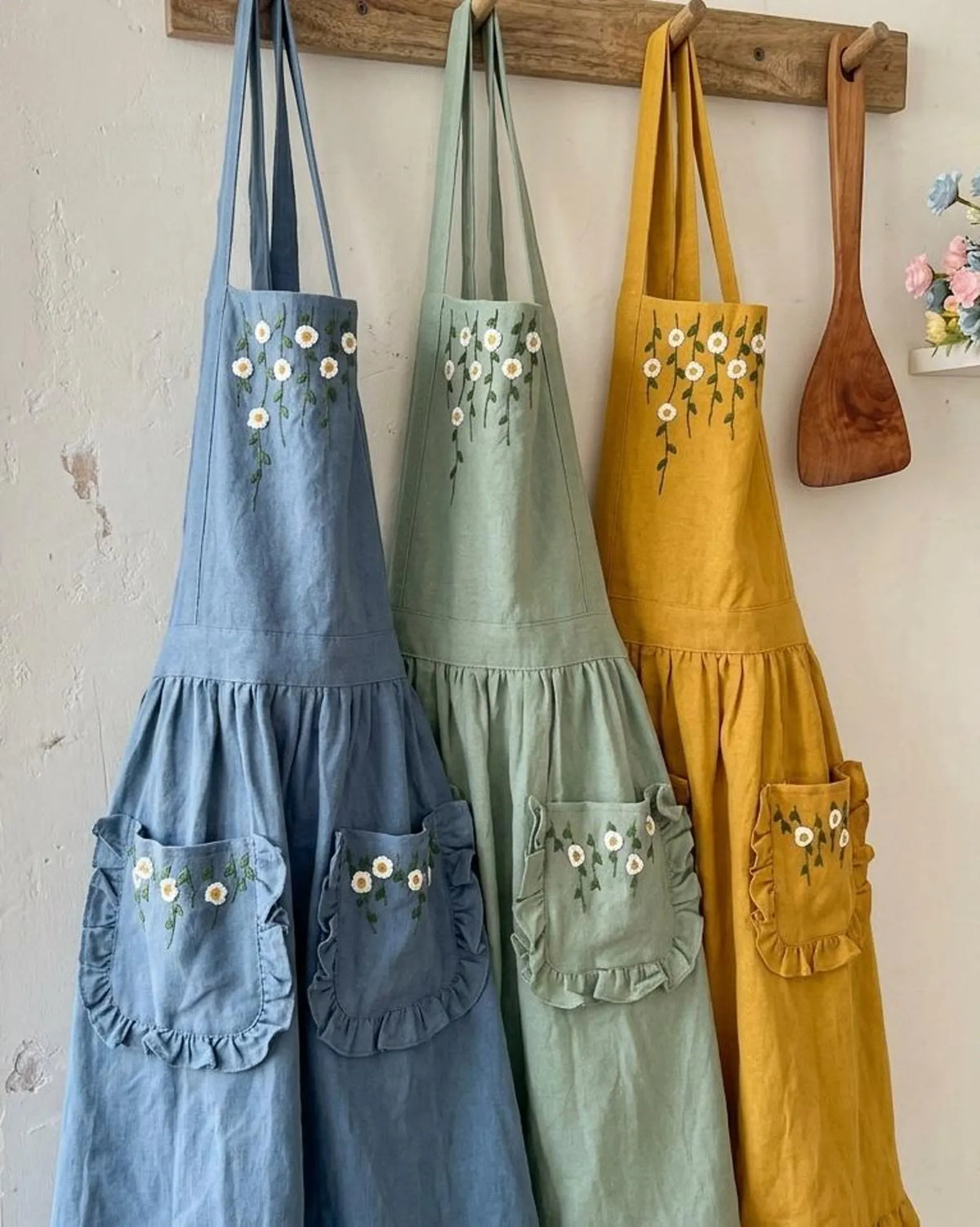 Three ruffled linen aprons in blue, green and yellow hanging side by side — hand-embroidered by The Thread Nest