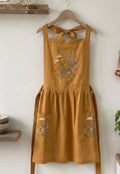A mustard linen apron on a wooden hanger with an embroidered wildflower bouquet — hand-embroidered by The Thread Nest