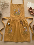 A mustard linen apron laid flat with an embroidered wildflower bouquet and two front pockets — hand-embroidered by The Thread Nest